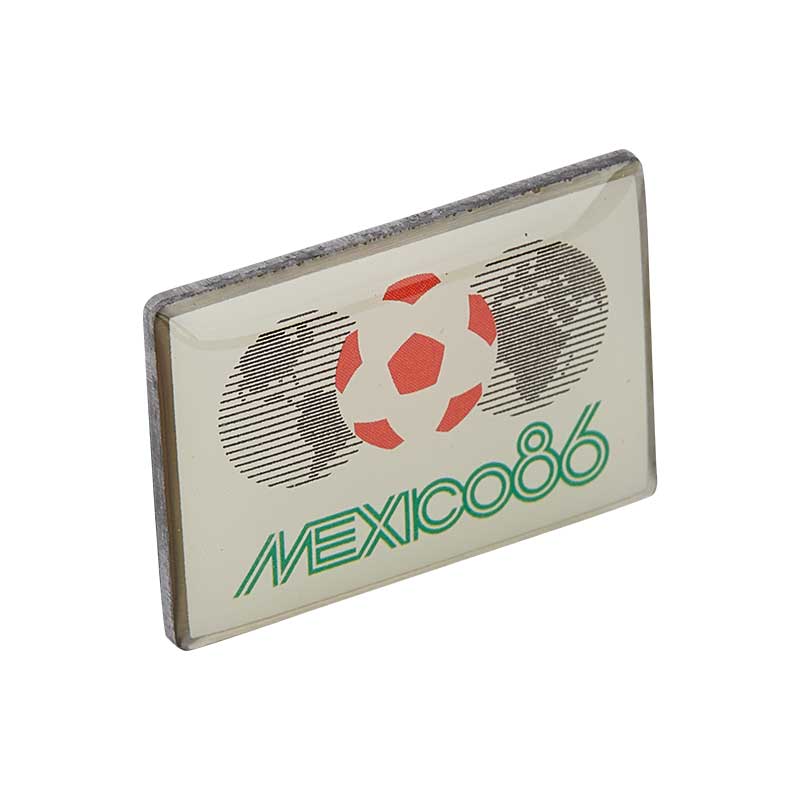 White pin with rounded corners, a white-red football in the center, and two globes on each side, representing the FWC in Mexico in 1986.
